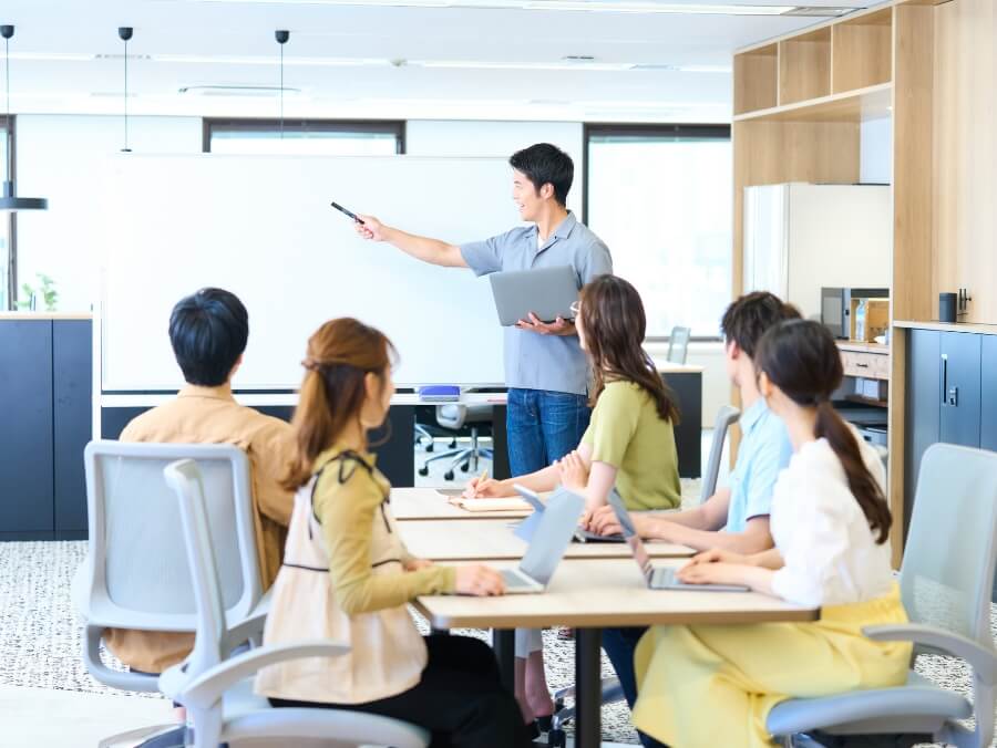 オフィス内で、一人の男性がホワイトボードの前に立ち、男性2人と女性3人の前で説明をしている
