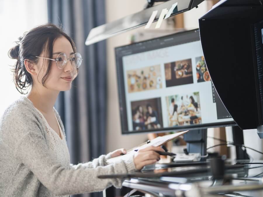一人の女性が左手にはバインダーを持ち、右手にはペンタブを持ち、パソコンのモニターを見ながら作業をしている