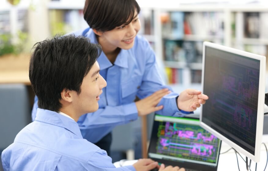 笑顔でいる男性と女性の2人の従業員が、外部モニターを見ながらネットワークシステムの稼働状況の確認をしている
